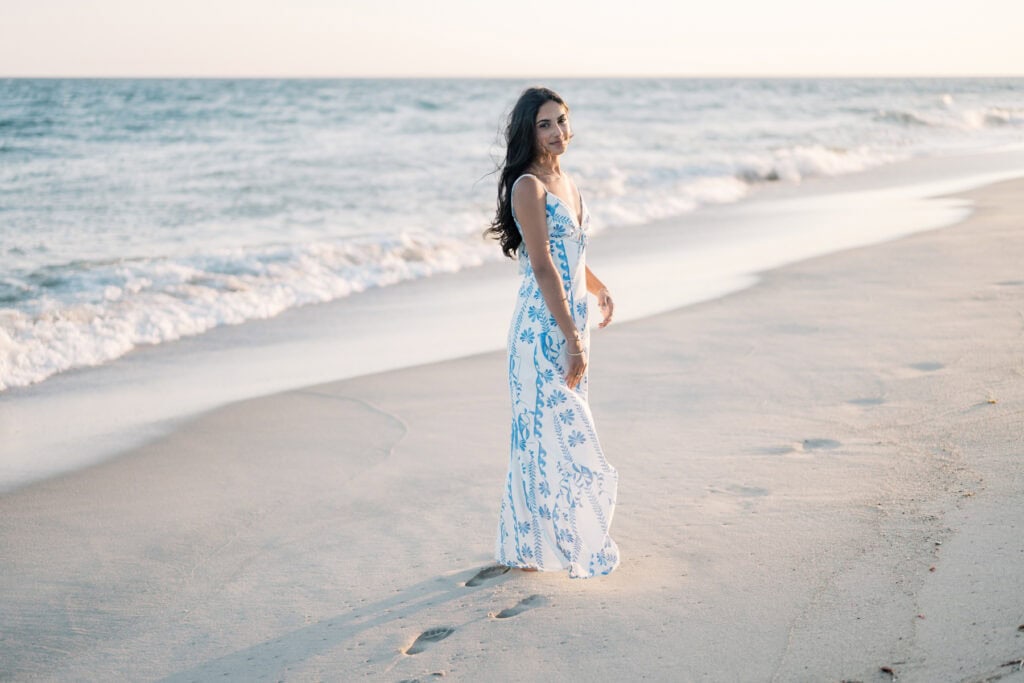 A woman in a white and blue dress stands on a sandy beach near the ocean, looking back and smiling as waves approach the shore.