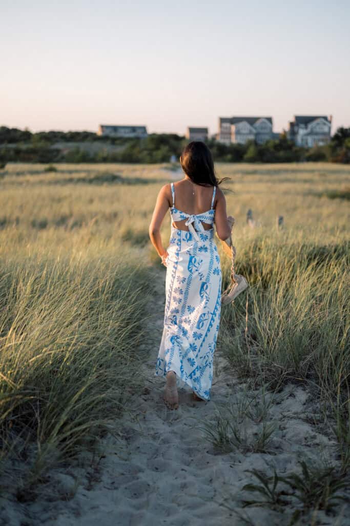 A woman in a blue and white floral dress walks barefoot through sandy grass towards distant houses at sunset, captured by photographer Kaity Farrell for Nantucket senior portraits.
