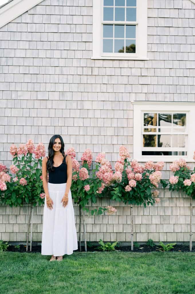 A woman in a black top and white skirt stands in front of a shingled house with pink hydrangeas along the wall, captured by photographer Kaity Farrell during a Nantucket summer senior session.