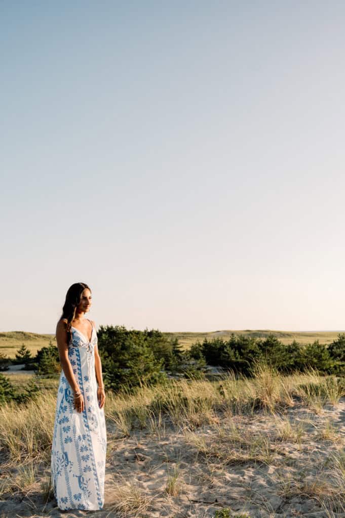 A woman in a white and blue sundress stands on sandy grass with trees and an open sky in the background.