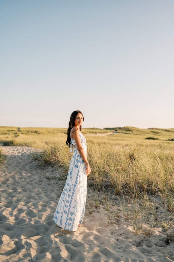 A woman in a white and blue patterned dress stands on a sandy path through grassy dunes under a clear sky, captured beautifully by photographer Kaity Farrell for Nantucket senior portraits.