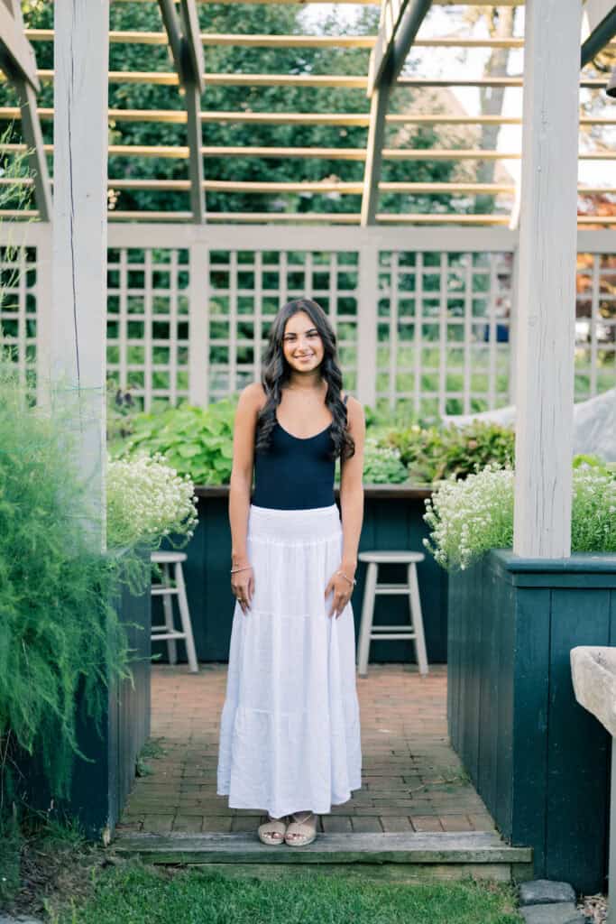 A woman in a black top and white skirt stands under a wooden pergola in a garden, smiling at the camera—captured beautifully by Nantucket senior portraits photographer Kaity Farrell.
