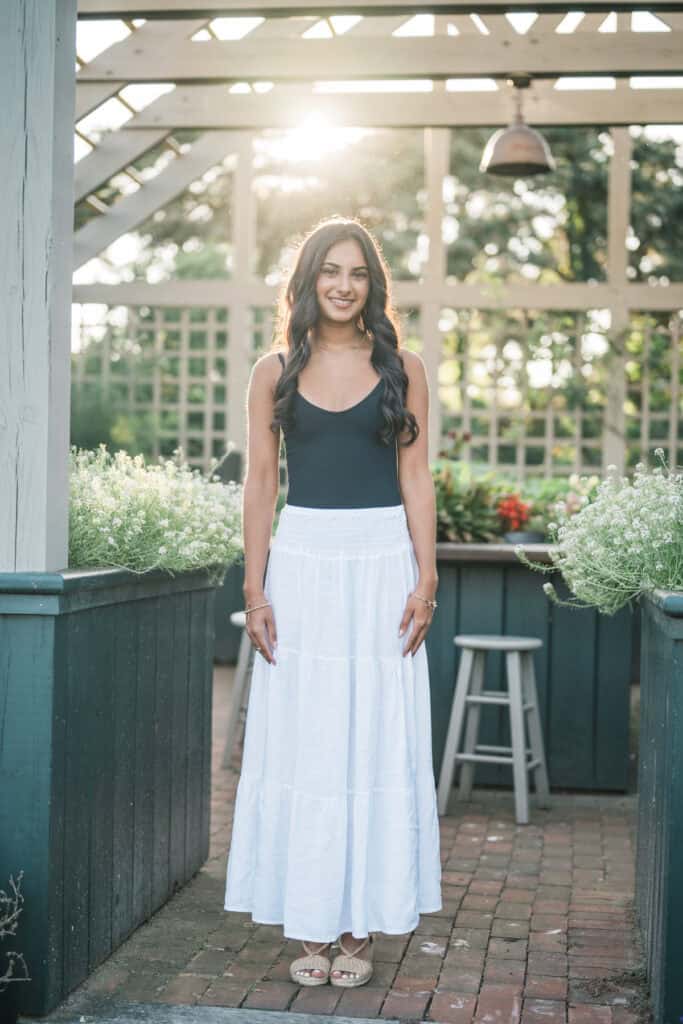 A young woman stands outdoors in front of a wooden trellis, wearing a black tank top and a long white skirt, sunlight shining behind her, beautifully captured by photographer Kaity Farrell for Nantucket senior portraits.