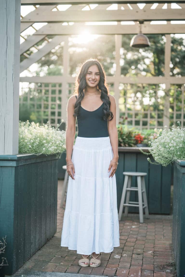 A young woman stands outdoors in front of a wooden trellis, wearing a black tank top and a long white skirt, sunlight shining behind her, beautifully captured by photographer Kaity Farrell for Nantucket senior portraits.