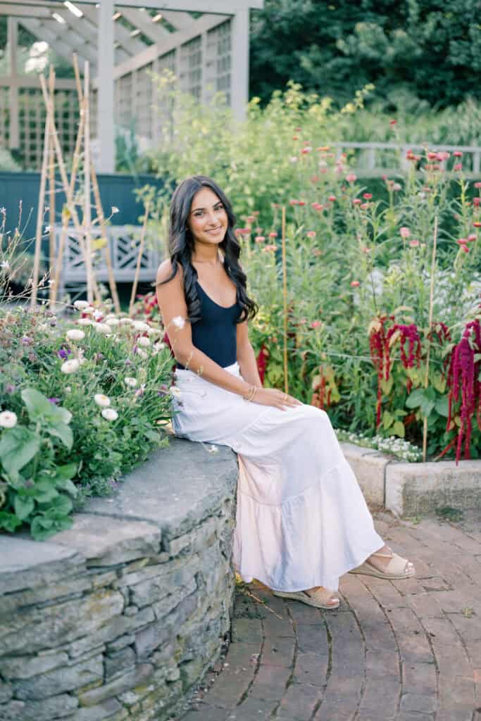 A woman with long dark hair, wearing a black top and white skirt, sits on a stone ledge in a garden surrounded by flowers and greenery—captured by photographer Kaity Farrell for Nantucket senior portraits.
