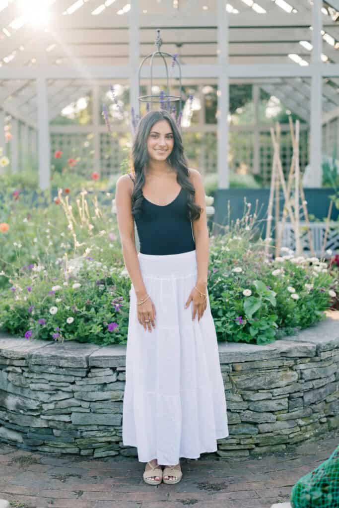 A woman stands in front of a stone flower bed, wearing a black top and long white skirt, in a sunlit garden setting—captured by photographer Kaity Farrell for Nantucket senior portraits.