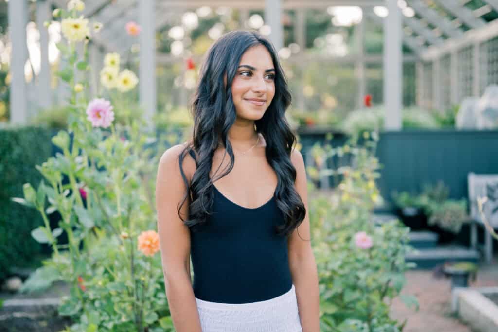 A woman with long dark hair wearing a black sleeveless top stands in a garden with blooming flowers and green plants, beautifully captured by Nantucket senior photographer Kaity Farrell.