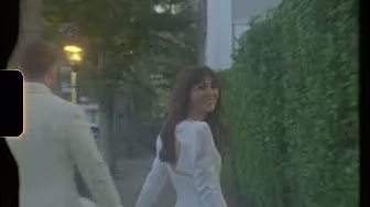 A woman in a white dress smiles while looking back, holding hands with a man in a light suit as they walk along a hedge-lined sidewalk.