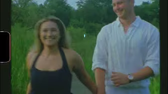 A woman and a man holding hands and walking outside on a path surrounded by greenery, both smiling.