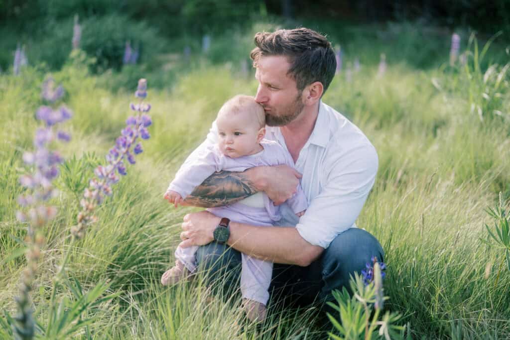 A man kneels in tall grass holding a baby in his arms, with wildflowers in the foreground and greenery in the background.
