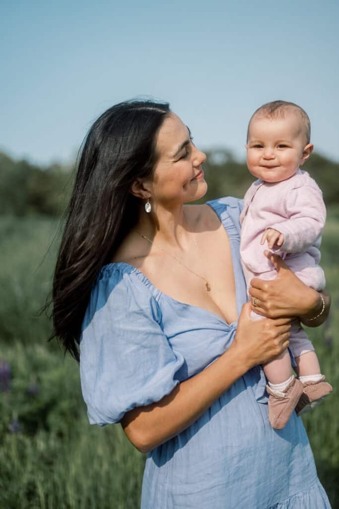 A woman in a blue dress holds a smiling baby in pink as they enjoy Nantucket family photos in a lupine field, captured by photographer Kaity Farrell beneath a clear sky.