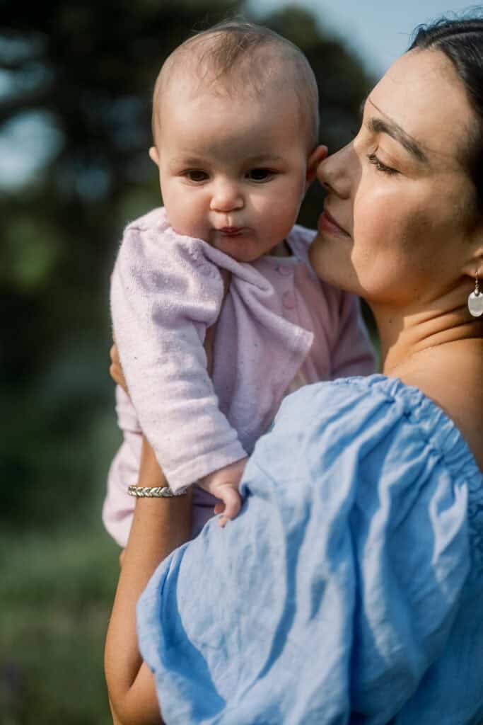 A woman in a blue off-shoulder top holds a baby dressed in pink outside, with greenery and trees softly blurred—capturing a tender moment during a Nantucket family portrait session by photographer Kaity Farrell.