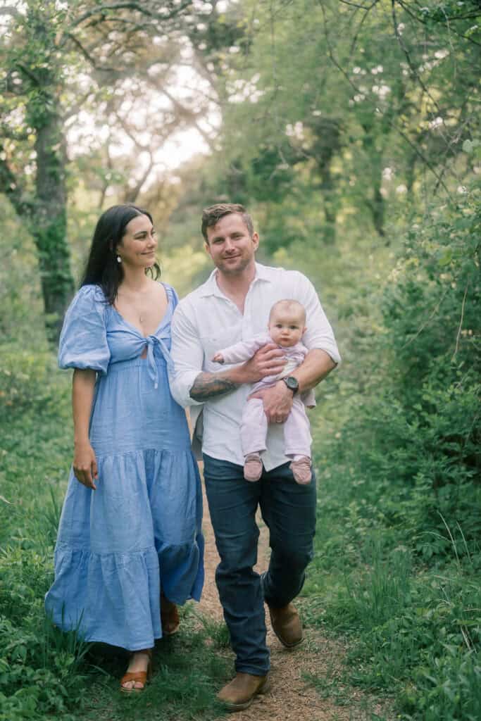 A man carries a baby while walking on a forest path beside a woman in a blue dress. The family is outdoors in a green, wooded area.