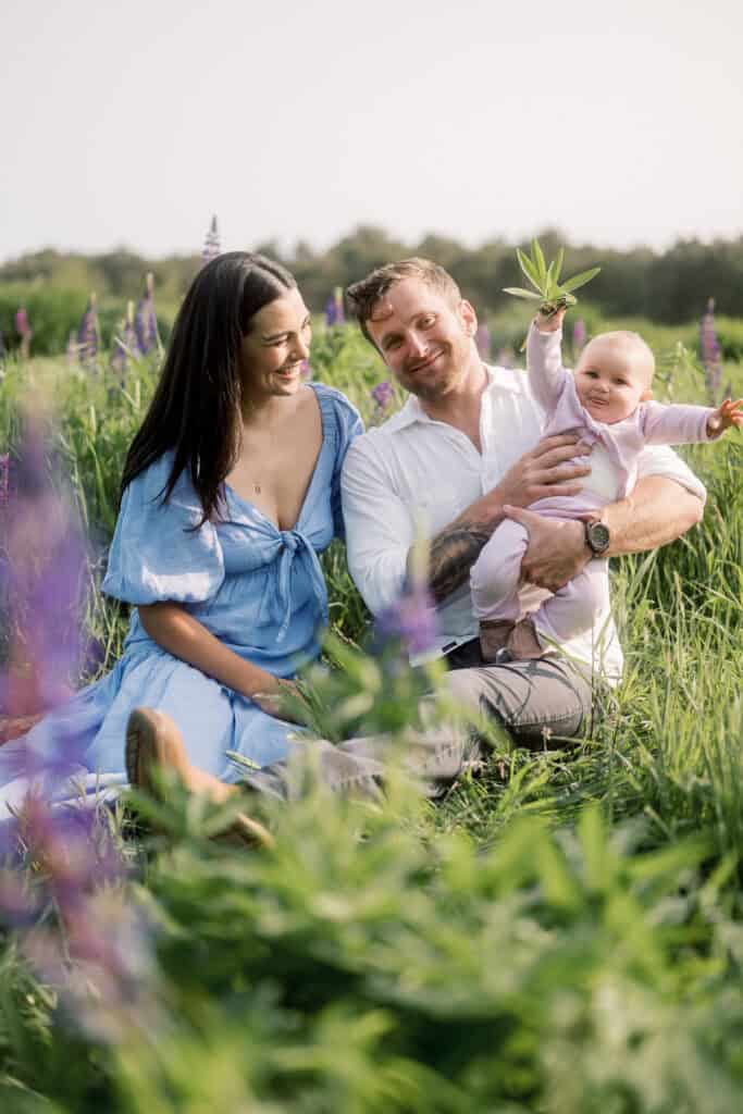 A man and woman sit on grass in a field with purple flowers, smiling at a baby the man is holding. The baby raises one arm while looking at the camera during a Nantucket family portrait session with photographer Kaity Farrell.