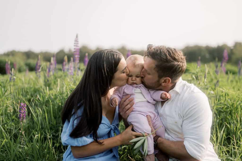 Two adults sit in a Nantucket flower field, surrounded by tall grass and purple blooms, holding and kissing a baby dressed in pink—a heartwarming moment captured by photographer Kaity Farrell.