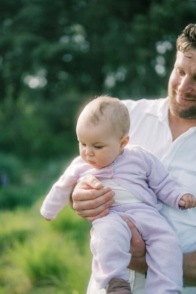 An father holding his baby daughter in a light purple outfit outdoors, surrounded by greenery—a moment captured by photographer Kaity Farrell, specializing in natural family photos on Nantucket.
