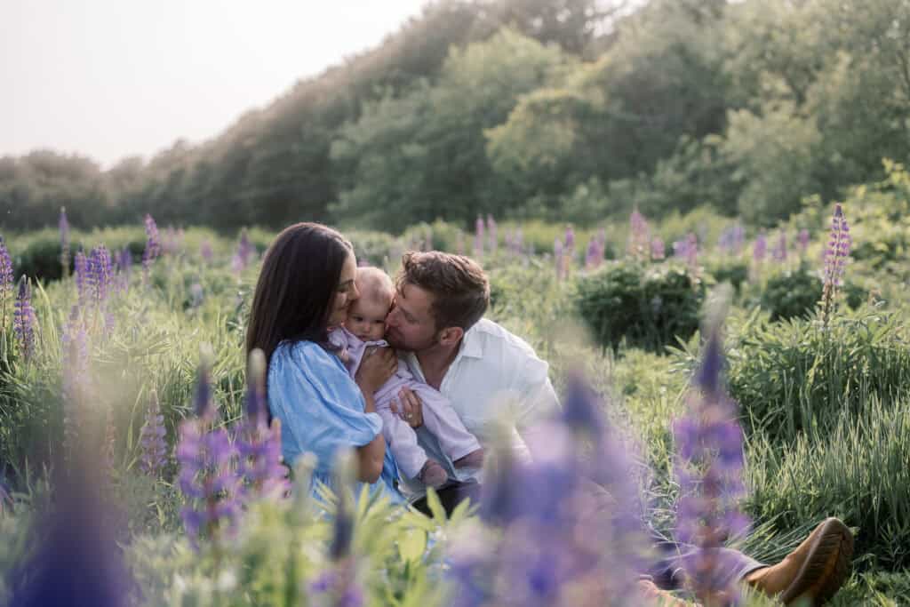 A couple sits in a field of purple flowers, holding and kissing a baby, surrounded by greenery and soft natural light—perfect for Nantucket family photos in lupine field by photographer Kaity Farrell.