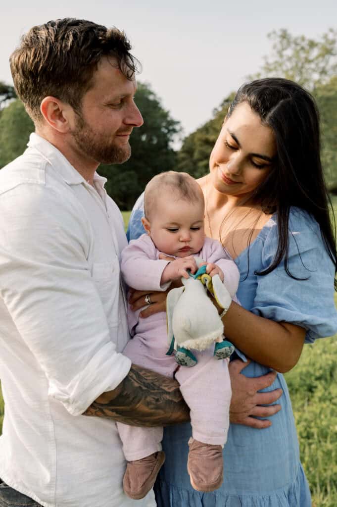 A man and woman stand outdoors holding a baby dressed in pink, who is holding a stuffed toy. Photographer Kaity Farrell captures this sweet moment during Nantucket spring family photos, with trees and grass in the background.