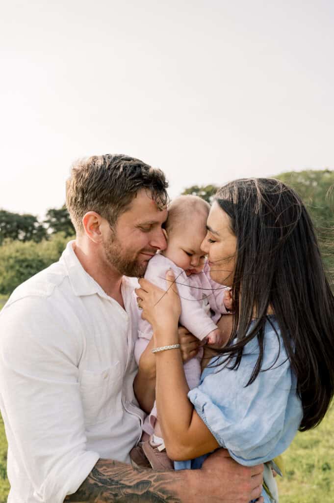 A man and woman stand outside in a lupine field holding a baby between them, all three touching heads affectionately—captured by photographer Kaity Farrell during Nantucket family photos.