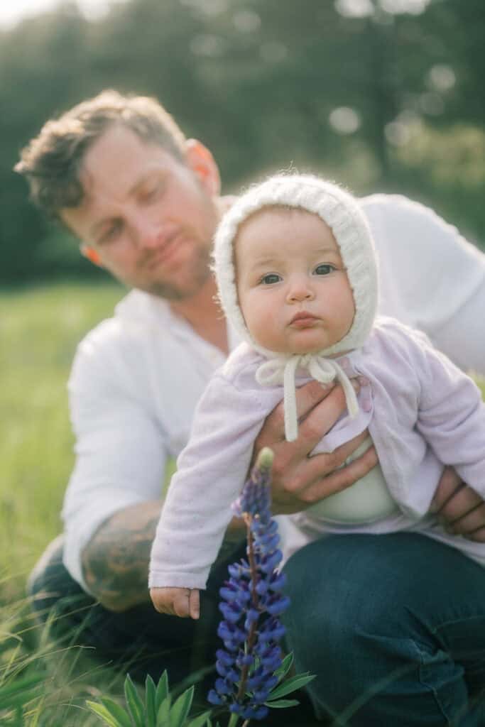 A man in a white shirt holds a baby in a knit bonnet and light sweater outdoors near purple wildflowers, with blurred greenery in the background—capturing sweet Nantucket spring family photos by photographer Kaity Farrell.