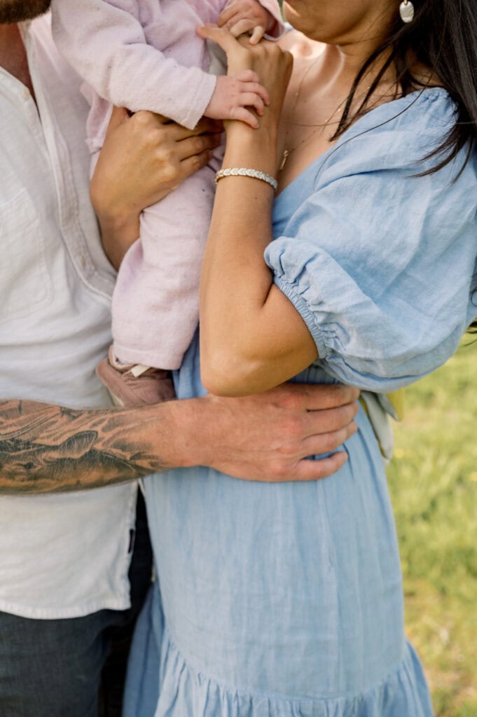 Three people stand close together; an adult in a blue dress holds a child in pink, while another adult with a tattooed arm embraces them during a Nantucket family portrait session with photographer Kaity Farrell.