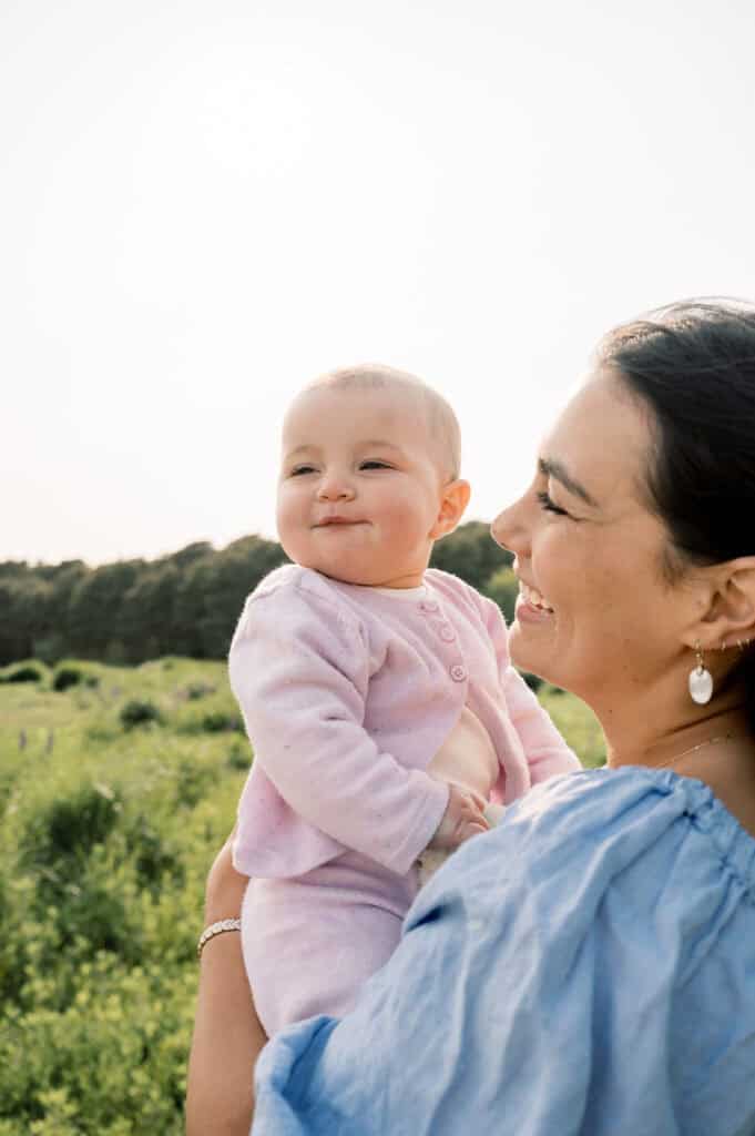 A mother in a blue blouse holds a smiling baby dressed in pink outdoors, surrounded by greenery and trees, beautifully captured by photographer Kaity Farrell specializing in Nantucket flower field portraits.