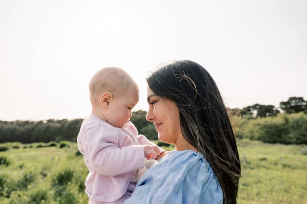 Woman holding a baby outdoors in a green field, both facing each other and smiling, with trees and a clear sky in the background—captured by Nantucket family photographer Kaity Farrell.