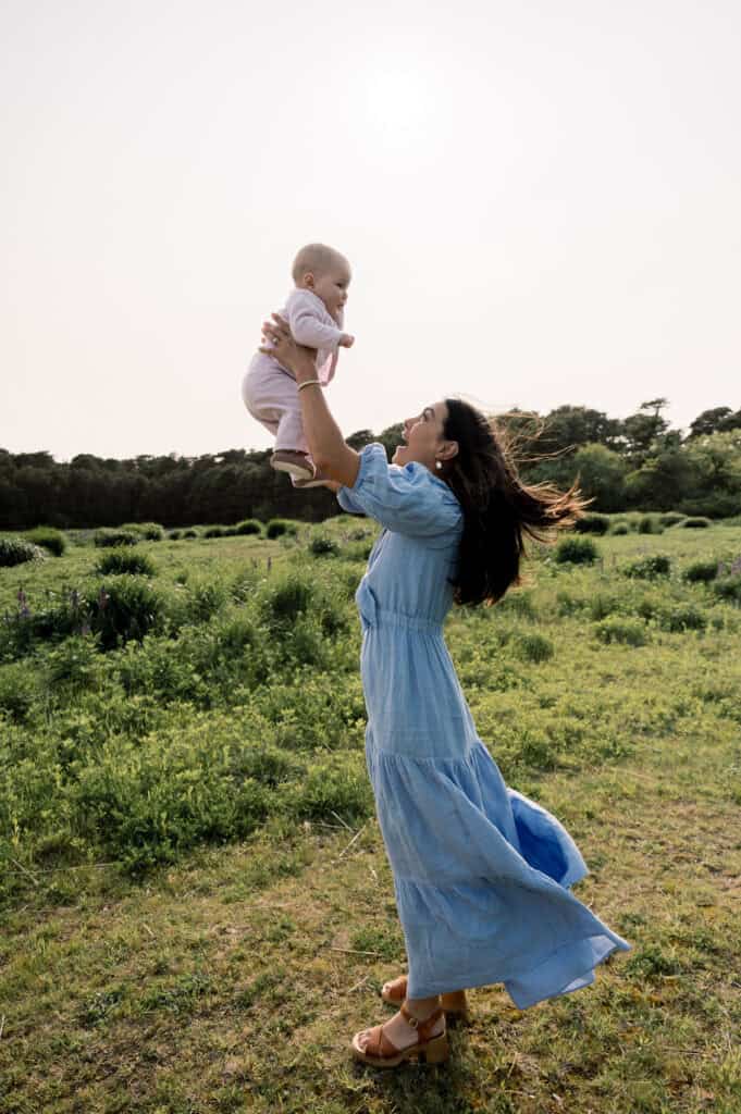A woman in a blue dress lifts a baby into the air while standing in a grassy field with trees behind them, capturing the joy of Nantucket outdoor family photos by photographer Kaity Farrell.