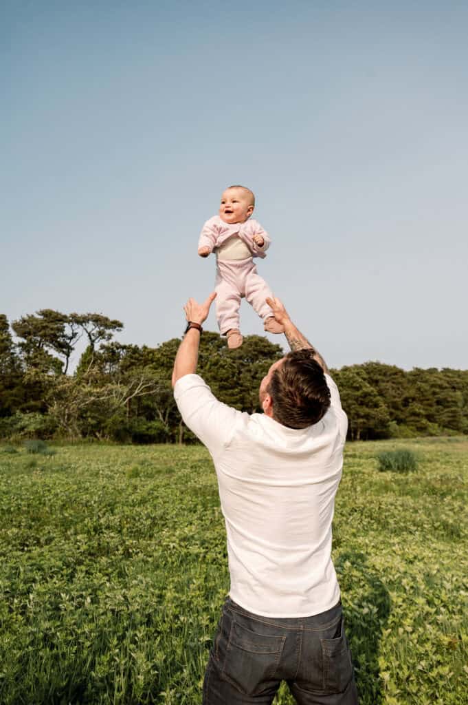 A father lifts his smiling baby girl into the air outdoors, standing in a grassy field with trees and a clear sky—the perfect setting for natural family photos Nantucket photographer Kaity Farrell is known for capturing.