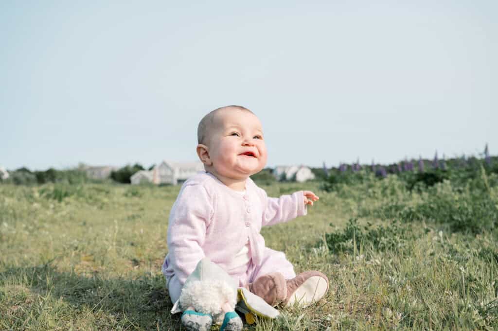 A baby wearing a light pink outfit sits on grass in a field, smiling, with houses and flowers visible in the background—captured by photographer Kaity Farrell during a Nantucket outdoor family photos session.