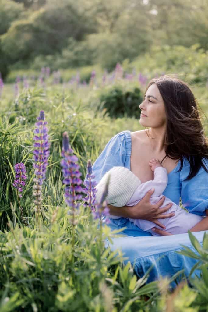 A woman in a blue dress breastfeeds her baby in a white hat among tall purple wildflowers, captured by photographer Kaity Farrell as part of Nantucket family photos in a vibrant lupine field.