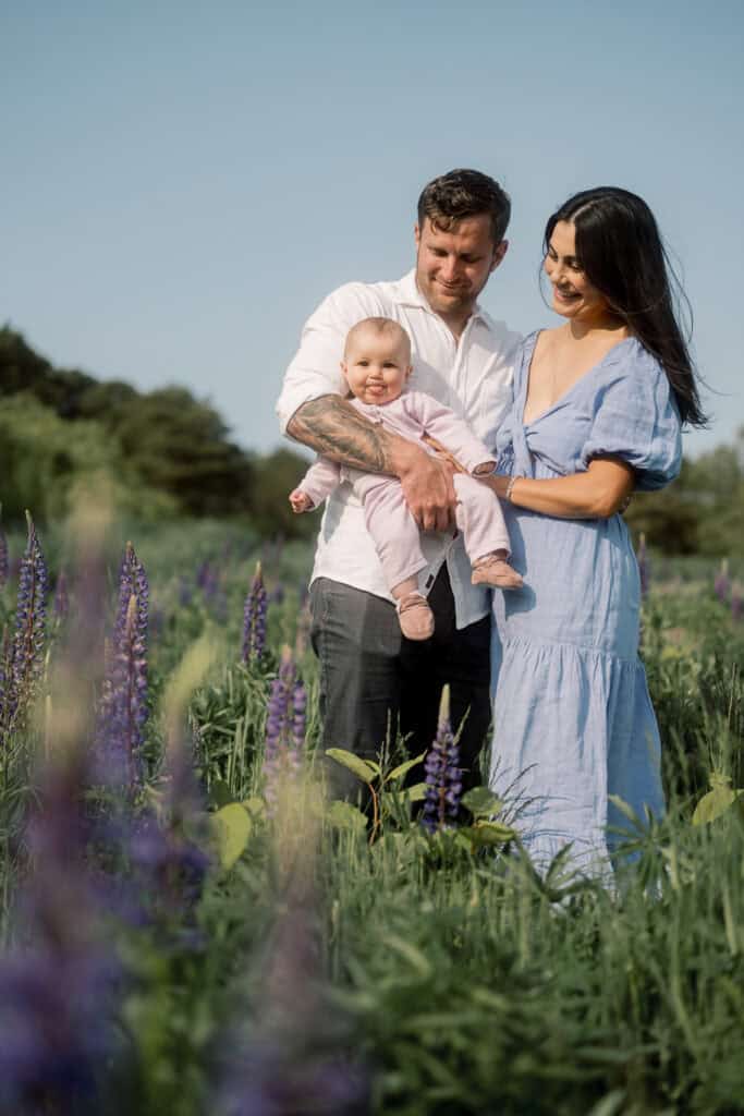 A man and woman stand in a field of purple flowers, smiling and holding a baby. Captured by photographer Kaity Farrell, these Nantucket flower field portraits feature a clear blue sky and trees in the background.