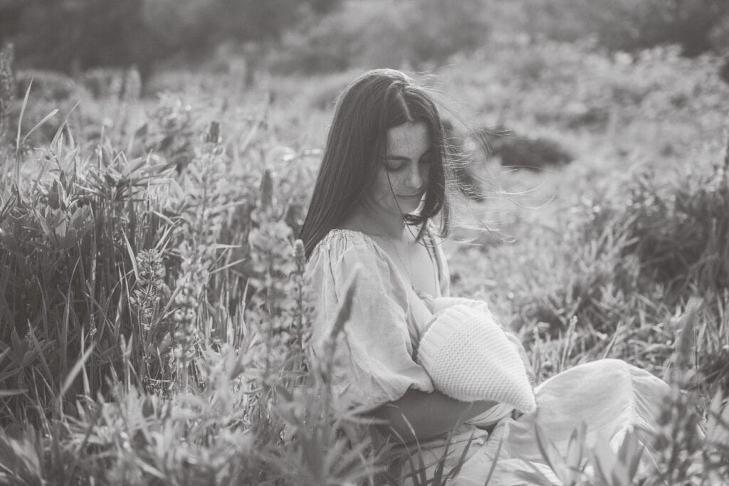 A woman sits in a grassy Nantucket flower field breastfeeding an infant wrapped in a blanket. Captured in black and white, this serene moment is part of photographer Kaity Farrell’s emotive portrait series.