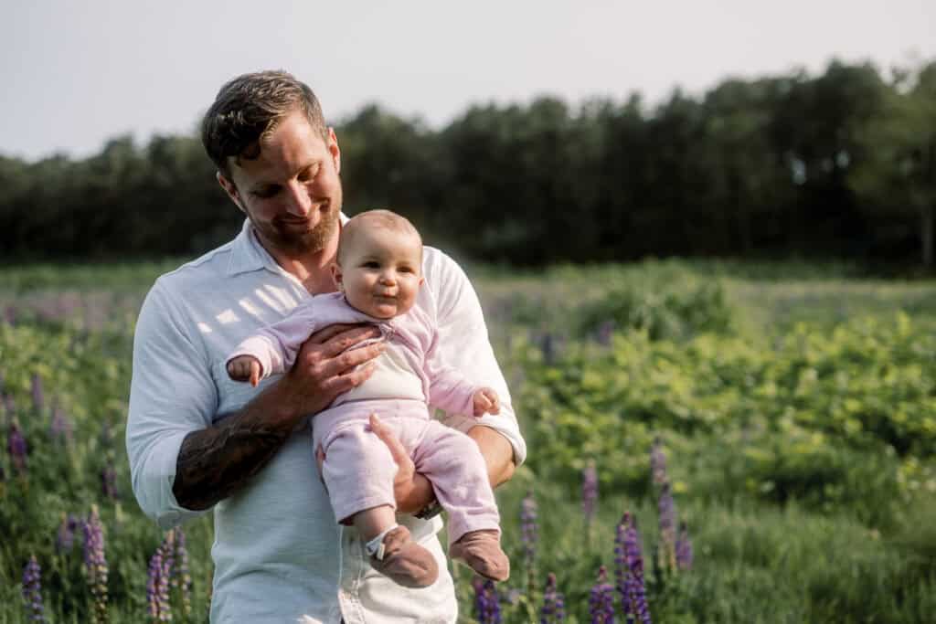 An adult holding a smiling baby outdoors in a field with greenery and purple flowers, trees in the background, captured by Nantucket family photographer Kaity Farrell.