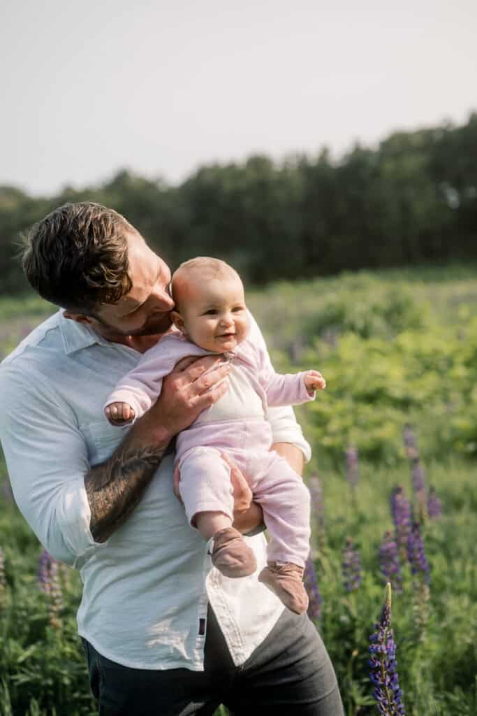 A man in a white shirt holds a smiling baby in a pink outfit in a field with purple wildflowers and greenery in the background.