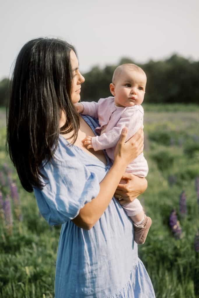 A woman in a blue dress holds a baby dressed in pink while standing in a field with green grass and purple flowers during a Nantucket family portrait session with photographer Kaity Farrell.