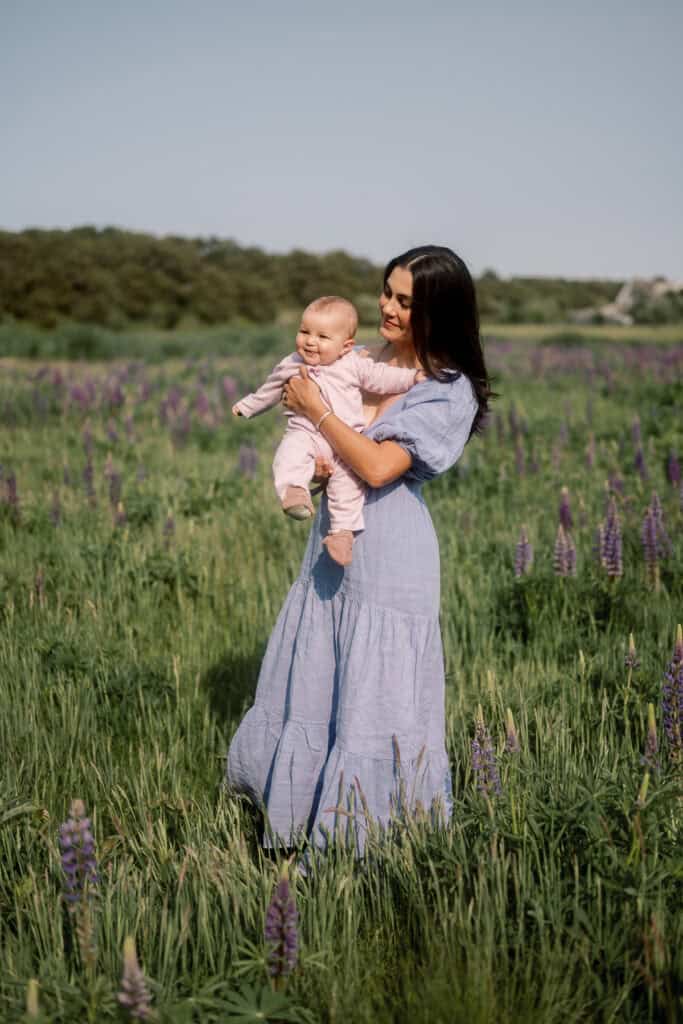 A woman in a light blue dress stands in a green field with purple flowers, holding a smiling baby dressed in pink—one of photographer Kaity Farrell’s beautiful Nantucket wildflower photos.