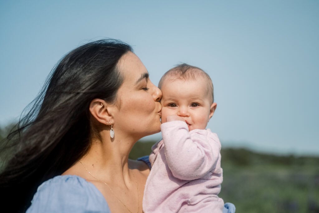Woman kissing a baby on the cheek while holding them outdoors; both are looking content with a clear sky in the background, beautifully captured by photographer Kaity Farrell during Nantucket family photos in a lupine field.